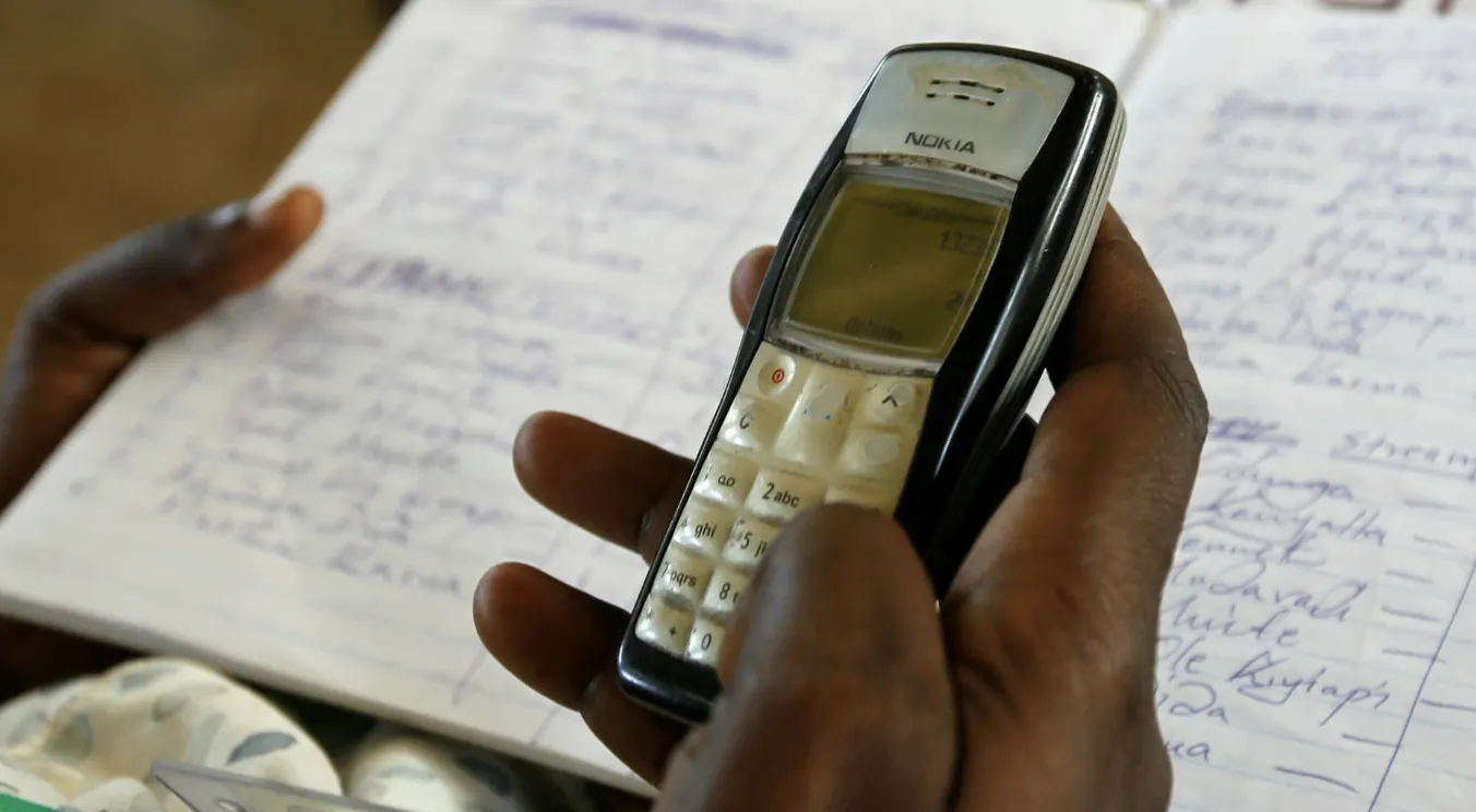 A hand holding a basic mobile phone over a handwritten paper register